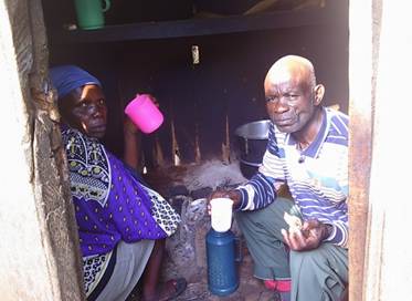 Two adults sit inside a small room near a hearth, holding cups, with cooking containers and household items visible around them.