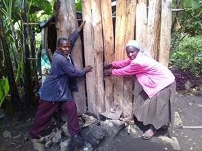 Two adults press wooden planks against the wall of a small structure made of vertical timber, reinforcing the exterior of the building in a rural setting.