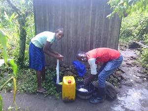 Two adults collect water from a pipe into containers placed on the ground beside a small wooden structure.
