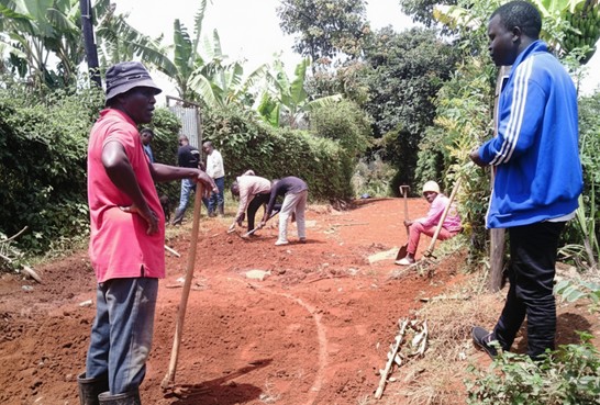 Adults stand and work along a red dirt road using hand tools, with greenery lining both sides of the path.