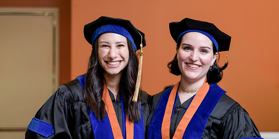Two PhD graduates smile in doctoral caps and gowns.