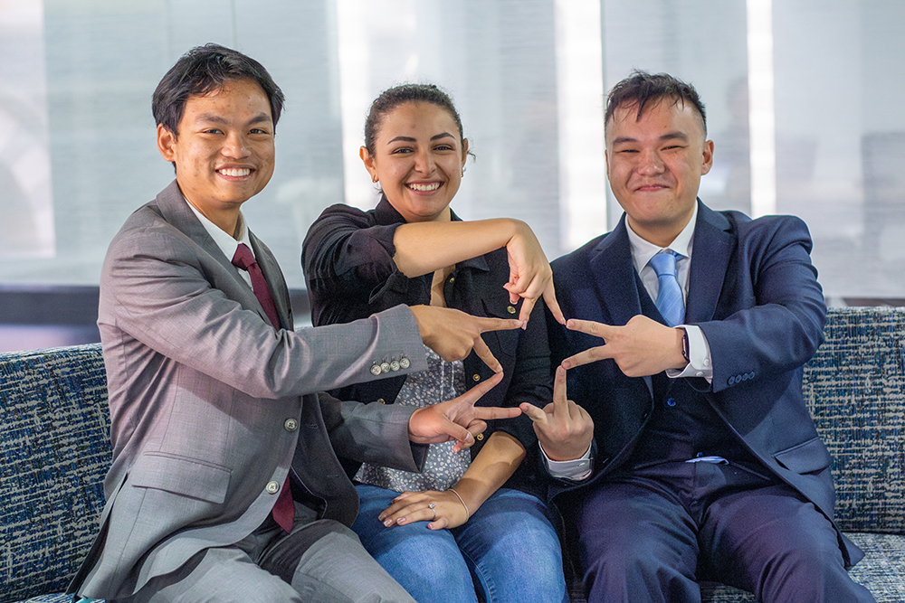 Three students in professional attire seated on a couch forming a star with their fingers.