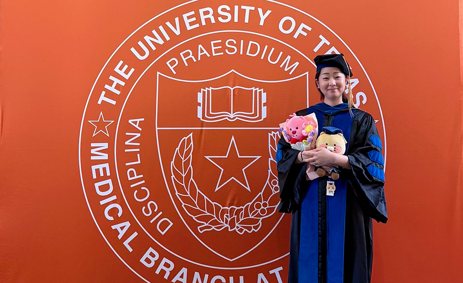 A doctoral graduate holds flowers and gifts while posing in academic regalia before the UTMB seal.