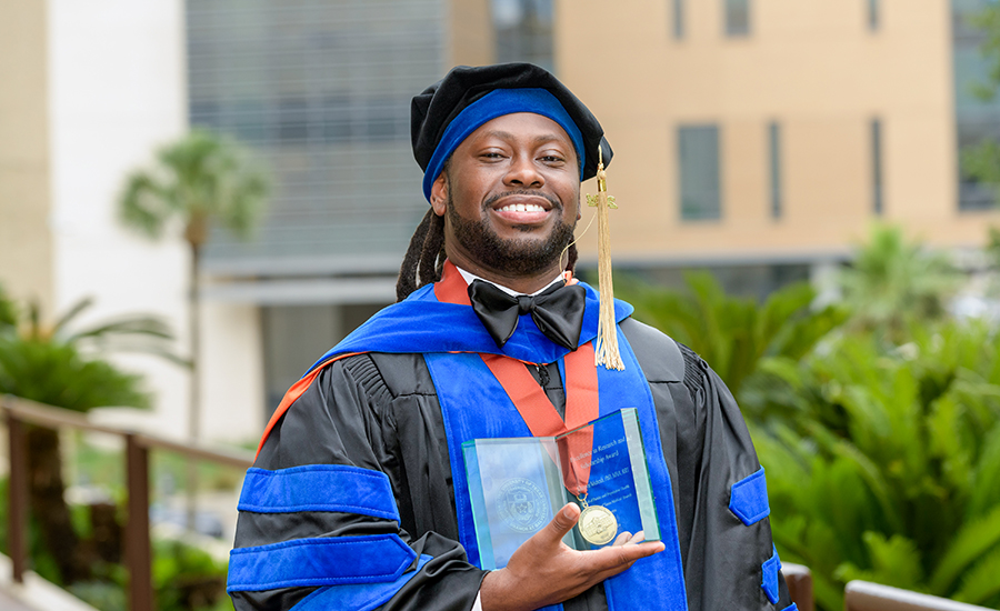 A Rehabilitation and Health Services Research PhD graduate poses in doctoral regalia while holding an award.