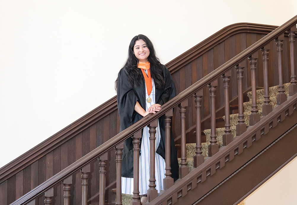 Klarisa Lopez smiles in graduation regalia and a medal while standing on a dark wooden staircase.