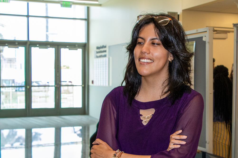 Monserrat smiles with her arms crossed in a university building while wearing a purple blouse.