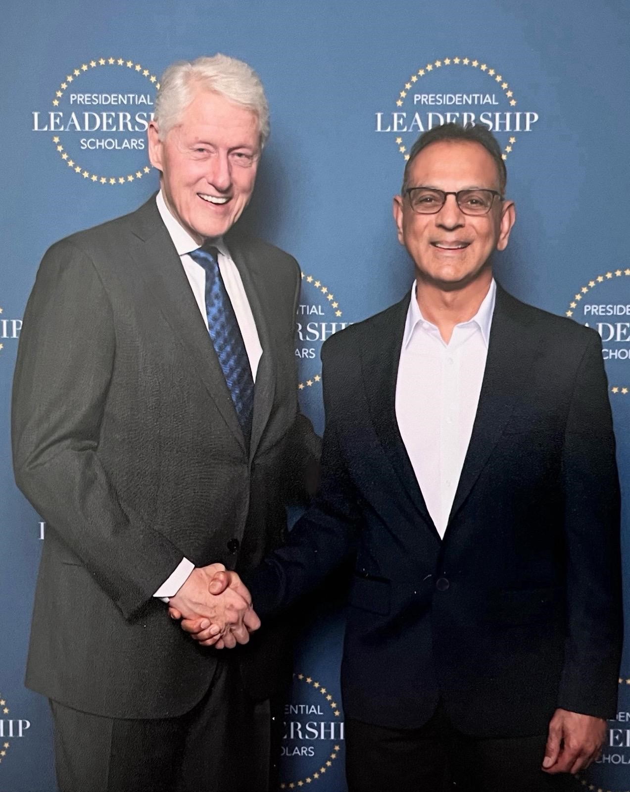 Dr. Suresh Bhavnani shaking hands with President Bill Clinton in front of a Presidential Leadership Scholars backdrop.