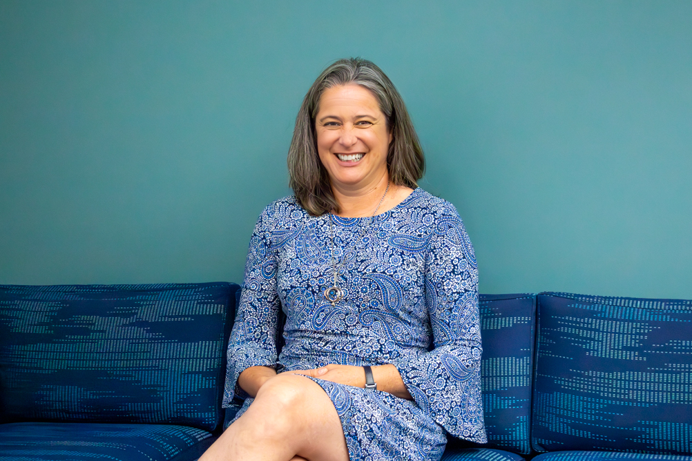 Dr. Heidi Spratt sits smiling on a blue bench against a teal wall, wearing a blue patterned dress and necklace