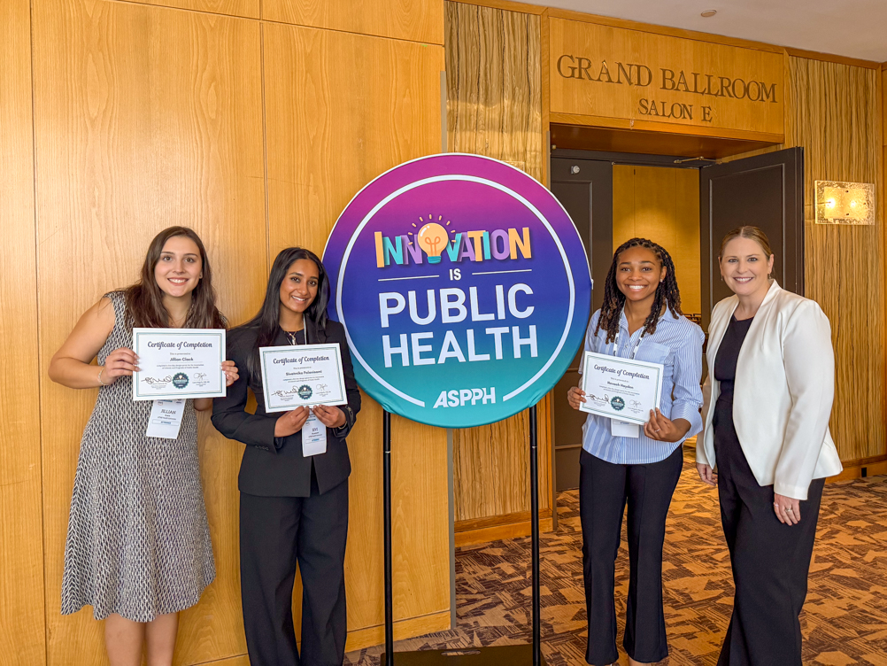 Jillian Clack, Sivi Palanisami, Nevaeh Hayden, and Innovation Lab facilitator Sarah Axelson hold certificates of completion next to the ASPPH Innovation is Public Health sign outside the Grand Ballroom at the Crystal Gateway Marriott.