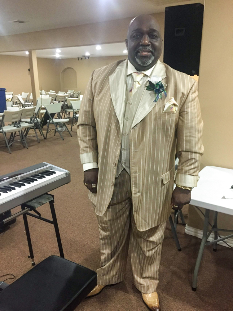 John Cooks stands beside an electronic keyboard in a tan pinstripe suit.