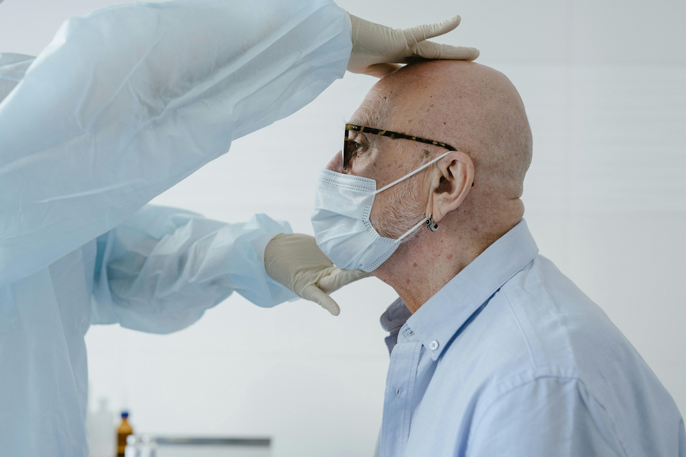 Clinician in protective gown and gloves examines an older man wearing glasses and a face mask during a medical visit.