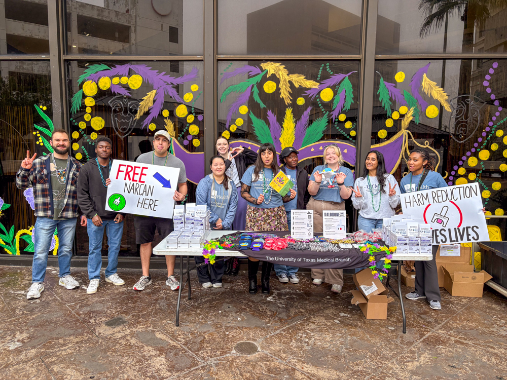 Student Association volunteers standing behind a table with Narcan distribution materials and signs reading Free Narcan Here and Harm Reduction Saves Lives during Mardi Gras