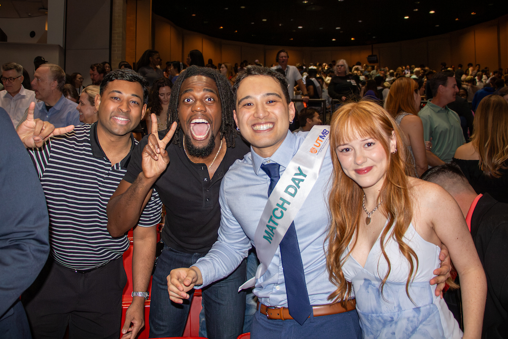 Four people smile and pose together in a crowded auditorium during Match Day