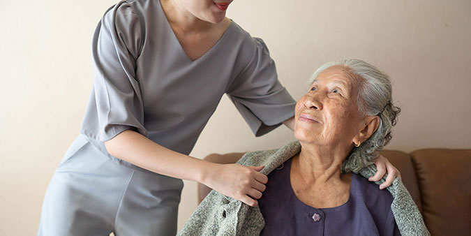 A medical staff member places a blanket on the shoulders of an elderly woman, who is looking up with a soft smile on her face