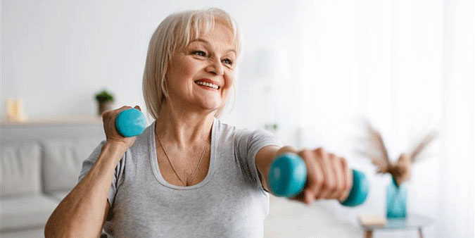 A woman in her 60s with blonde hair performs a workout with two small dumbbells in her hands