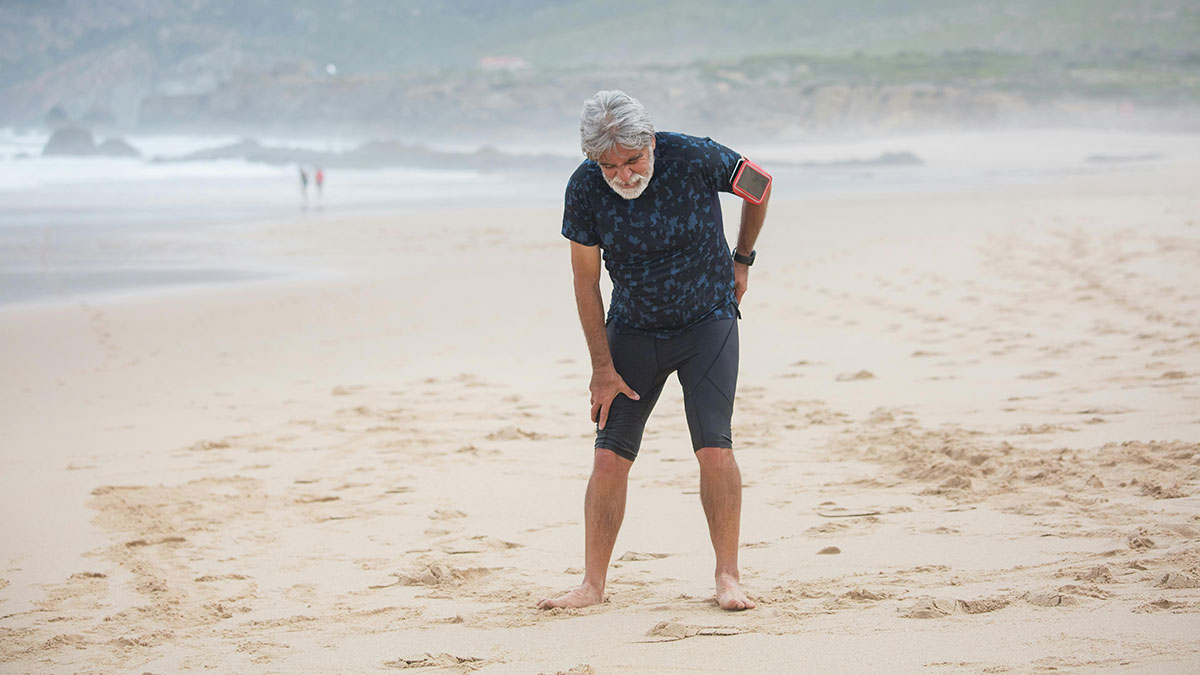 A gray-haired man on a beach bends over feeling his lower back