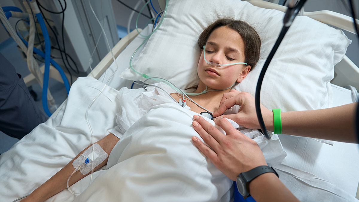 A child lies in a hospital bed with her eyes closed and tubes attached to her body as someone checks her heart