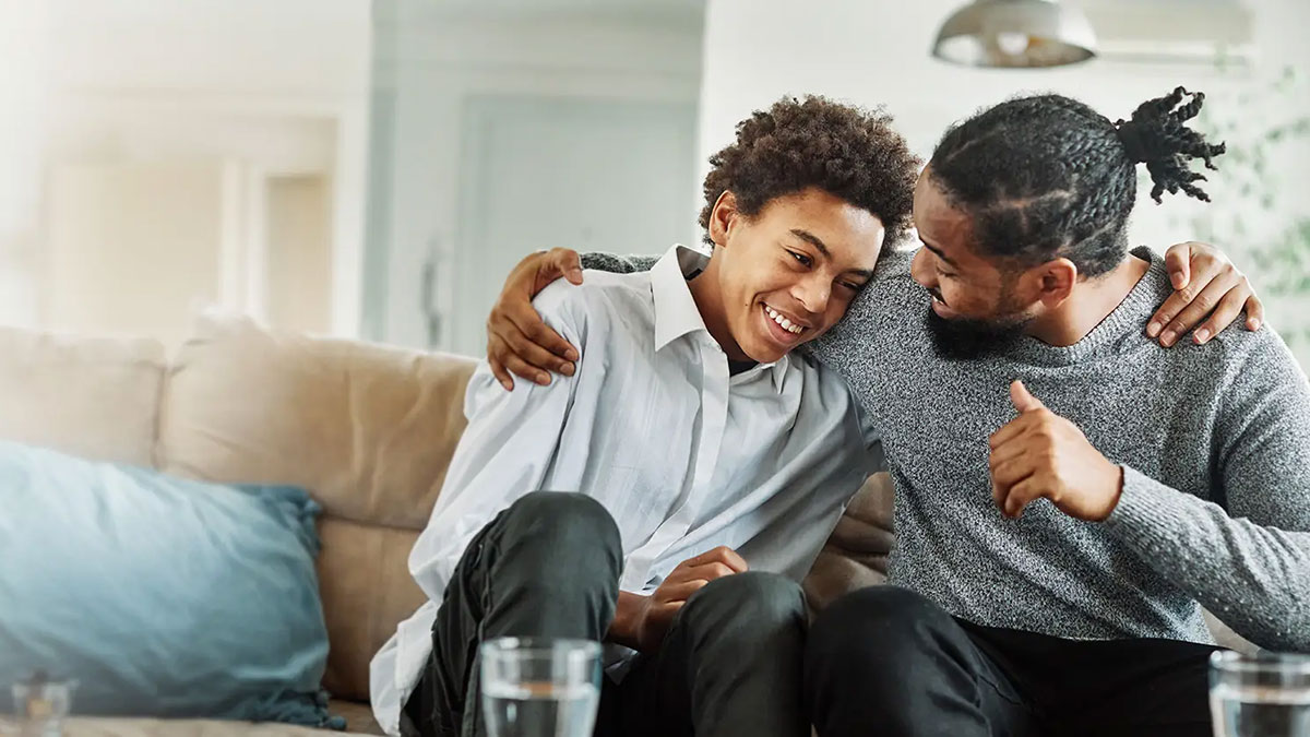A teen boy sits on the sofa, smiling, a his dad puts an arm around his shoulders