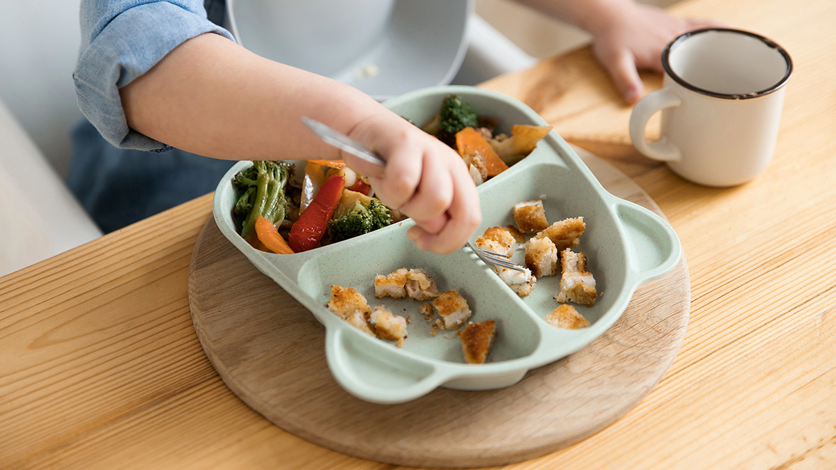 A child's hands are seen holding a fork over an animal-shaped food dish with chicken and vegetables.