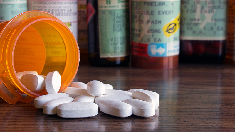 White pills are spilled from an open bottle, with additional prescription pill bottles upright in the background.