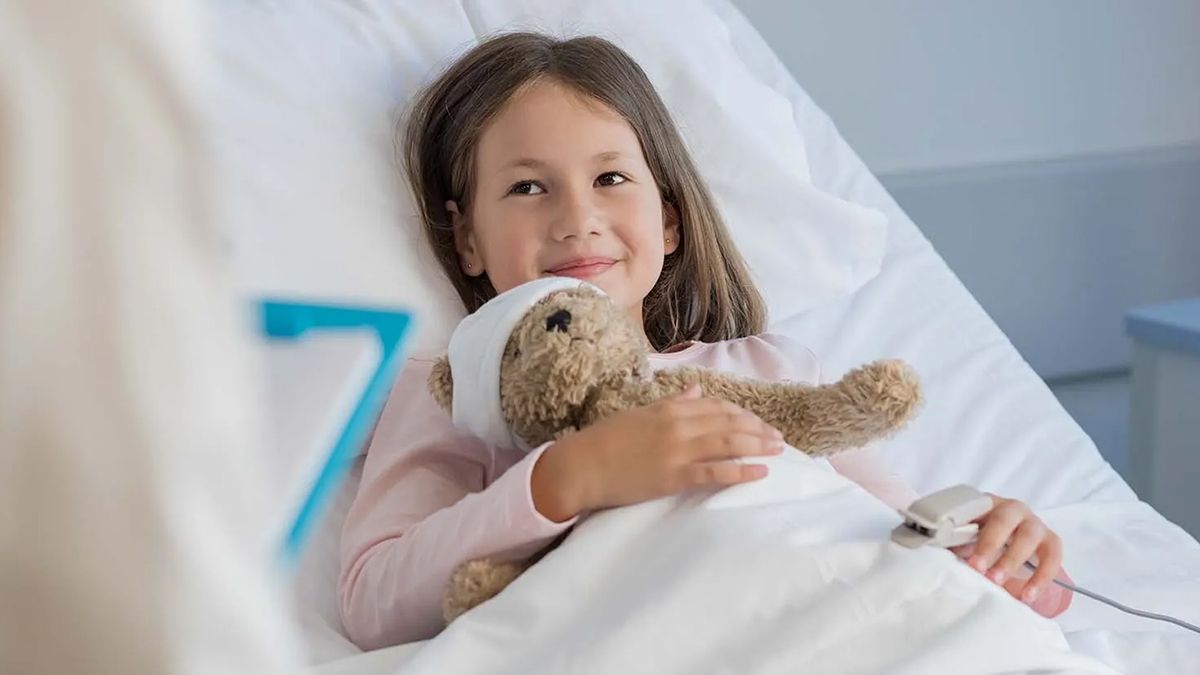 A young girl lies in a hospital bed holding a teddy bear