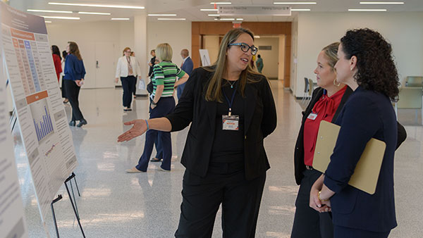 A woman with long blonde hair wearing a black suit shows her poster to two session attendees.
