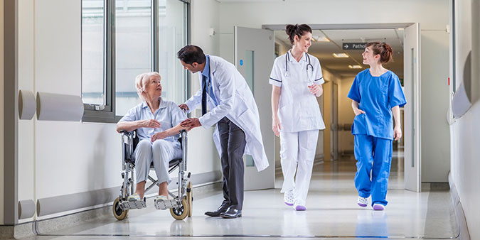 A male doctor assists a senior woman in a wheelchair in a hospital hallway as two other clinicians walk by