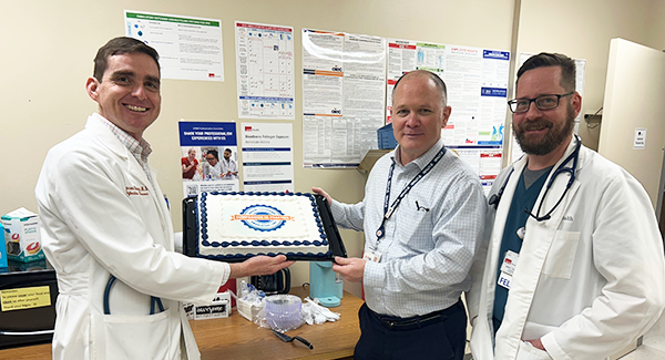 Dr. Lucas Blanton and Dr. Miguel Cabada pose, smiling and holding a rectangular cake with the Compassion in Practice logo. Dr. Jason Lauderdale stands next to them.