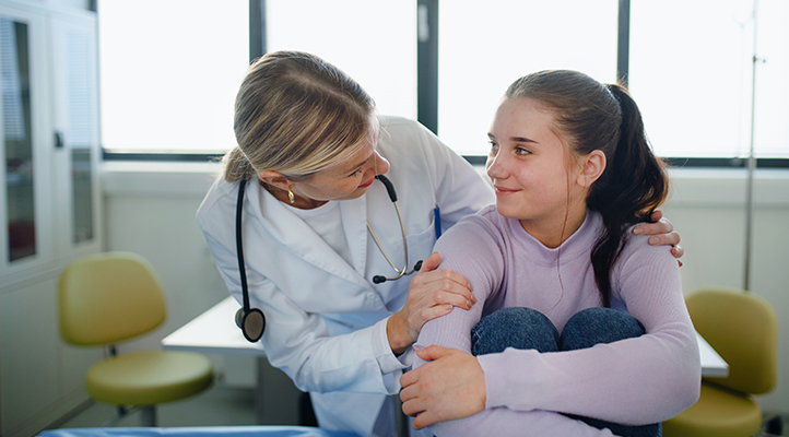 A woman health care provider in a white coat places her hands on a teen girl's shoulders; they are smiling at each other.