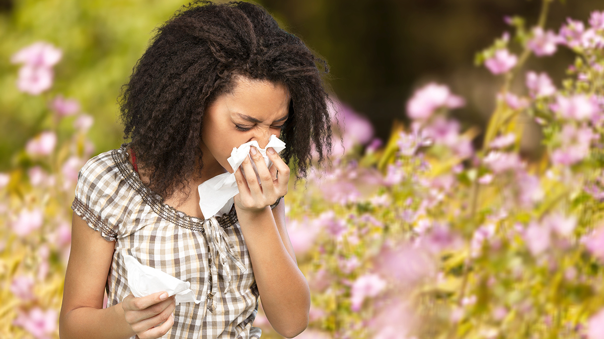 A woman standing in a field of flowers is sneezing into a tissue.