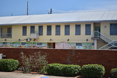 A yellow building with several windows on the second floor and a colorful mural on the first floor, behind a brick half wall.