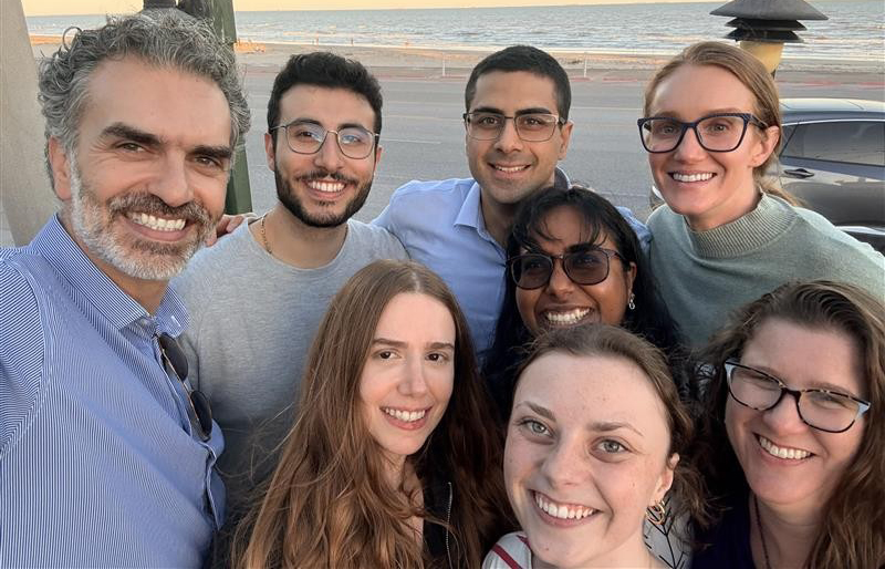 A group selfie of eight smiling team members posing together outdoors with the ocean beach in the background