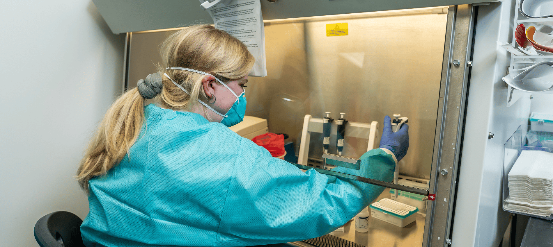 Emily Edgar wears a lab coat and mask while she works in a biosafety cabinet