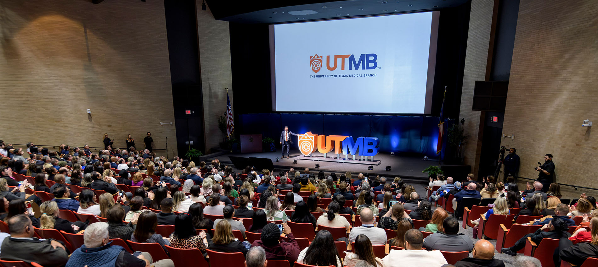 Audience seated in a large auditorium watching Dr. Reiser on stage at the University of Texas Medical Branch logo launch event, with a large screen and illuminated UTMB signage displayed behind him.