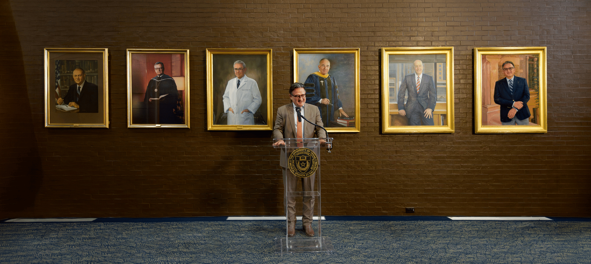 Dr. Reiser speaks at a podium in front of a wall displaying six painted portraits of past University of Texas Medical Branch presidents.