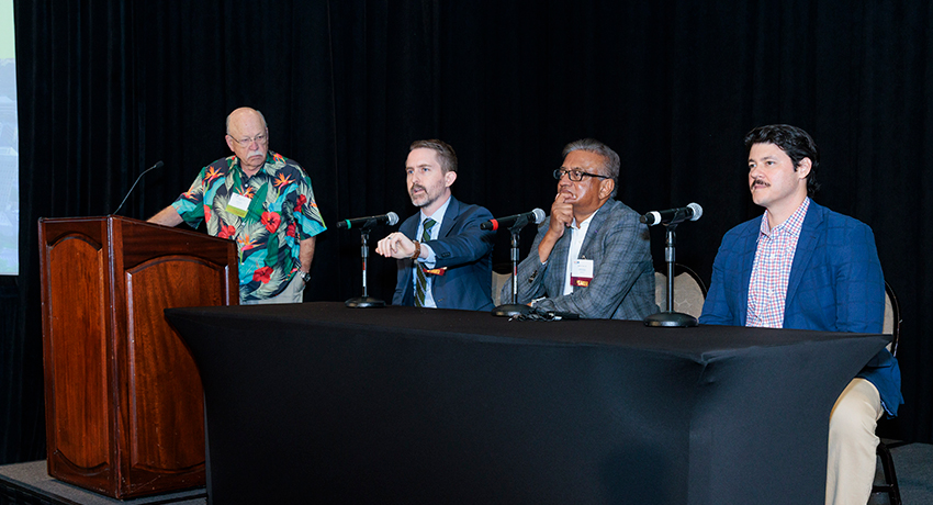 Three men in business attire sit at a long black table while a man in a colorful, tropical shirt stands at a podium during a panel discussion.