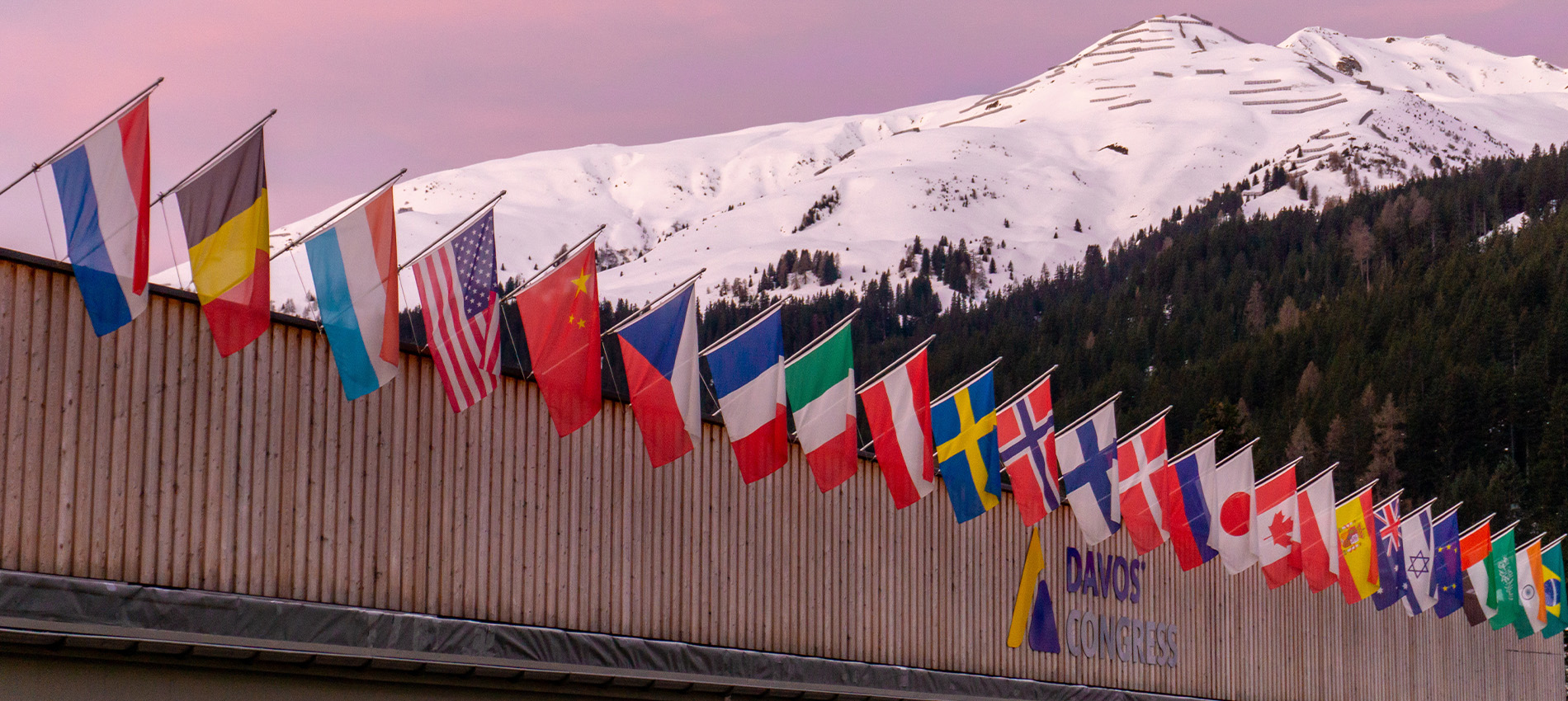 Row of international flags lining the Davos Congress Center with snow-covered mountains and a pink sky in the background.