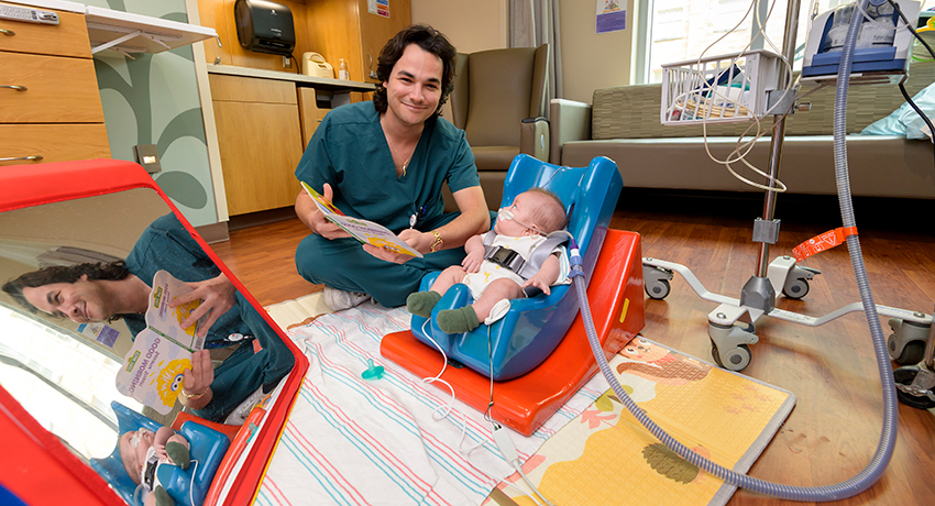 A man in scrubs sits on the floor in the NICU, reading a book to an infant who reclines in a special seat.