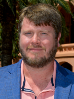  Headshot shows a man with brown hair and beard wearing a blue jacket and pink shirt.