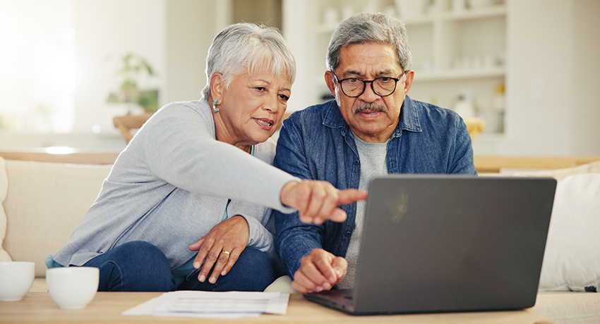 Two adults sit on a couch looking at a laptop, with one person pointing at the screen. Papers and two cups are on the table in front of them.