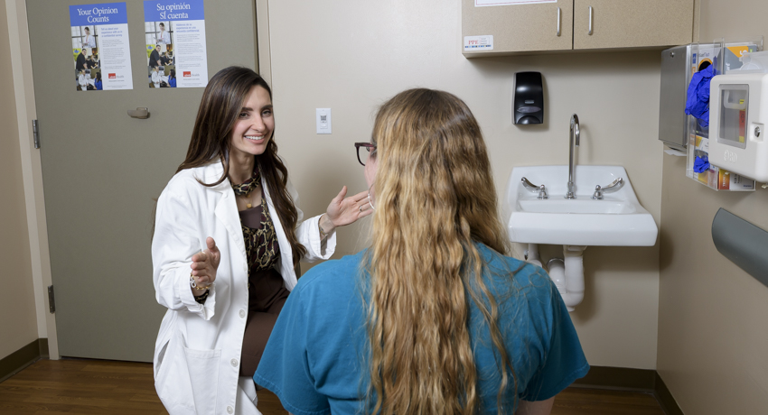 Health care provider in a white coat seated across from a patient in an exam room, speaking during a consultation. A sink, cabinets, and wall-mounted medical supply containers are visible in the room.