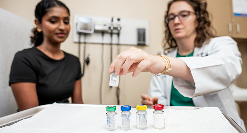 Clinician places a small vial on an exam table beside four color-coded vials while a seated patient looks on in an examination room.