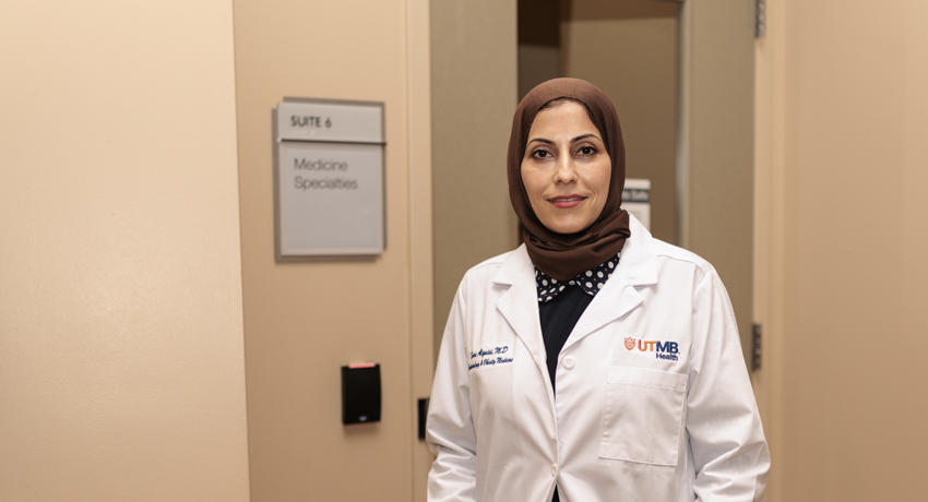 Dr. Sura Alqaisi stands in the hallway outside a UTMB Medicine Specialists clinic wearing a white lab coat. She faces the camera with a welcoming expression near the entrance to the clinic suite