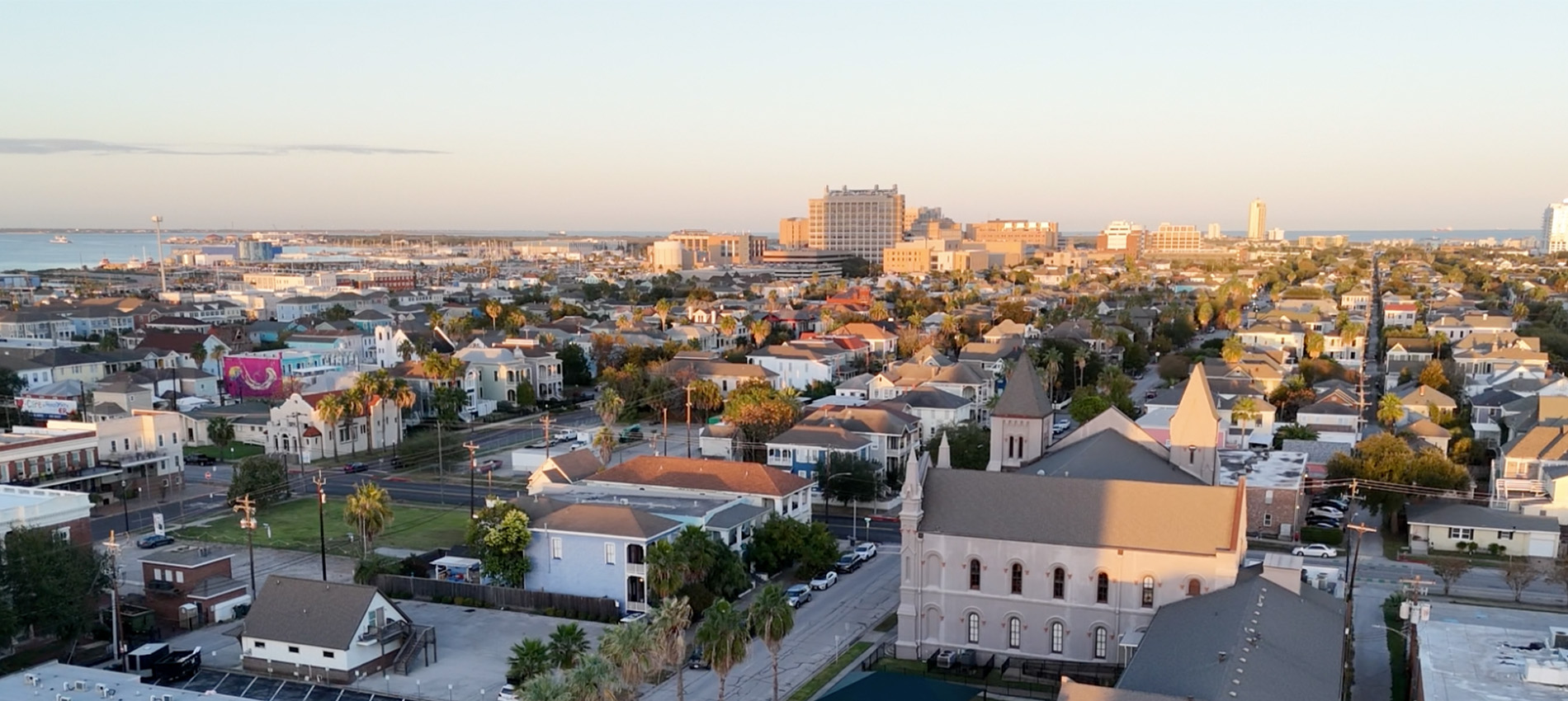 Aerial view of a coastal cityscape at sunset, showing residential blocks, tree‑lined streets, and larger buildings near the shoreline in the distance.