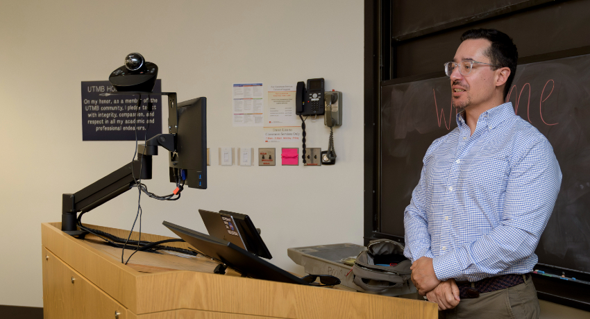 Dr. Alberto Aparicio stands at the front of a classroom, speaking to students during a bioethics lecture on longevity at the John Sealy School of Medicine at UTMB. A podium with computer monitors and a chalkboard are visible in the classroom.