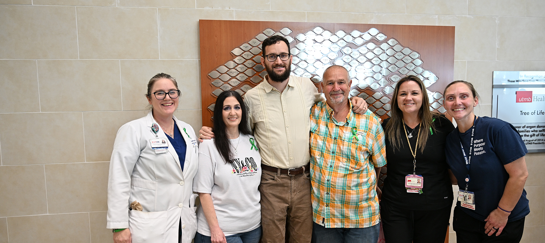 Four women and two men, some dressed in uniforms of health care workers, gather in front of a large hallway display of a tree with silver plaques as leaves.