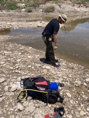 Austin Johnson works along the rocky shoreline as part of the massive search-and-rescue mission following the 2025 flooding in Kerr County. Person stands on a rocky shoreline beside shallow water, holding a rope or cable that extends into the water. Diving gear, including a tank and equipment bag, rests on the ground nearby.