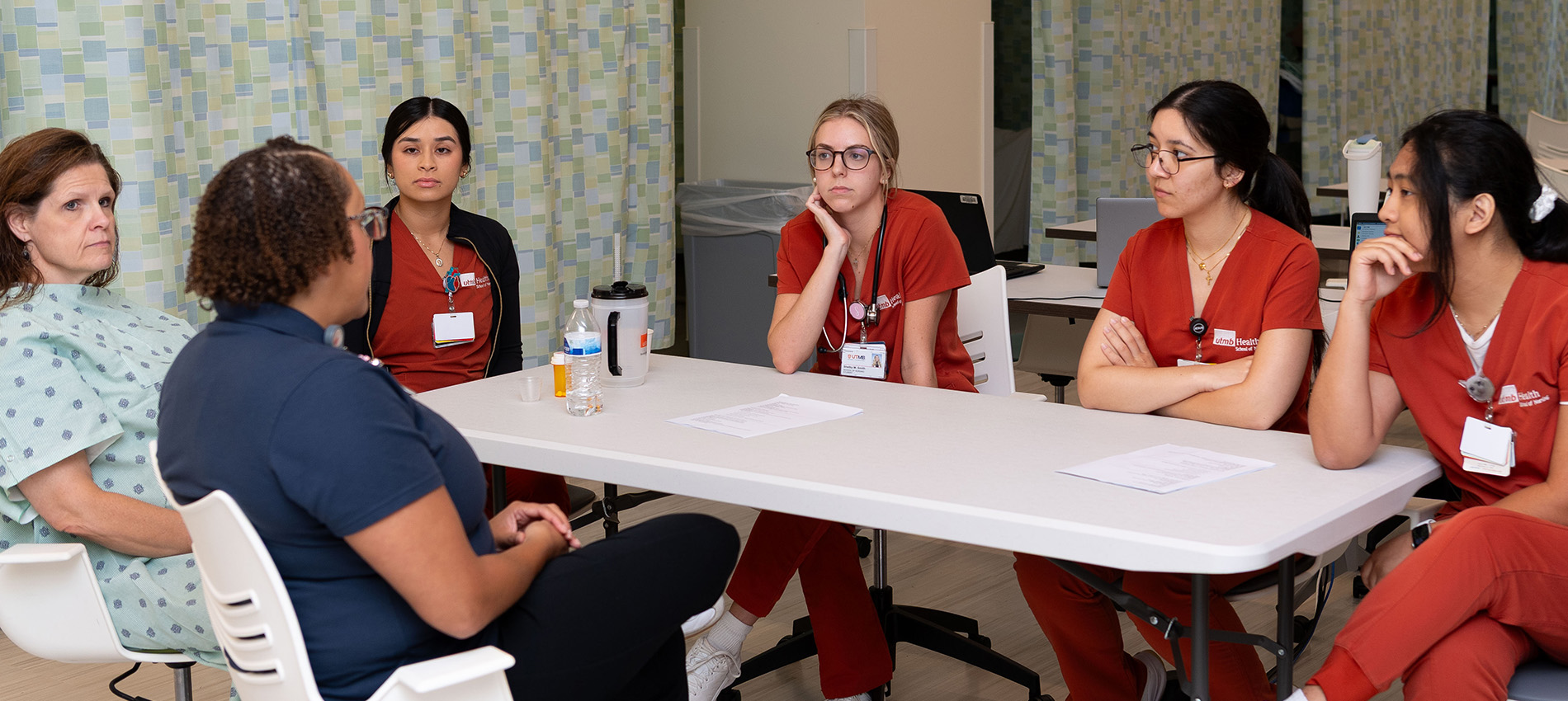 A nursing instructor, four nursing students and a standardized patient in a hospital gown gather around a table to discuss their simulation experience.