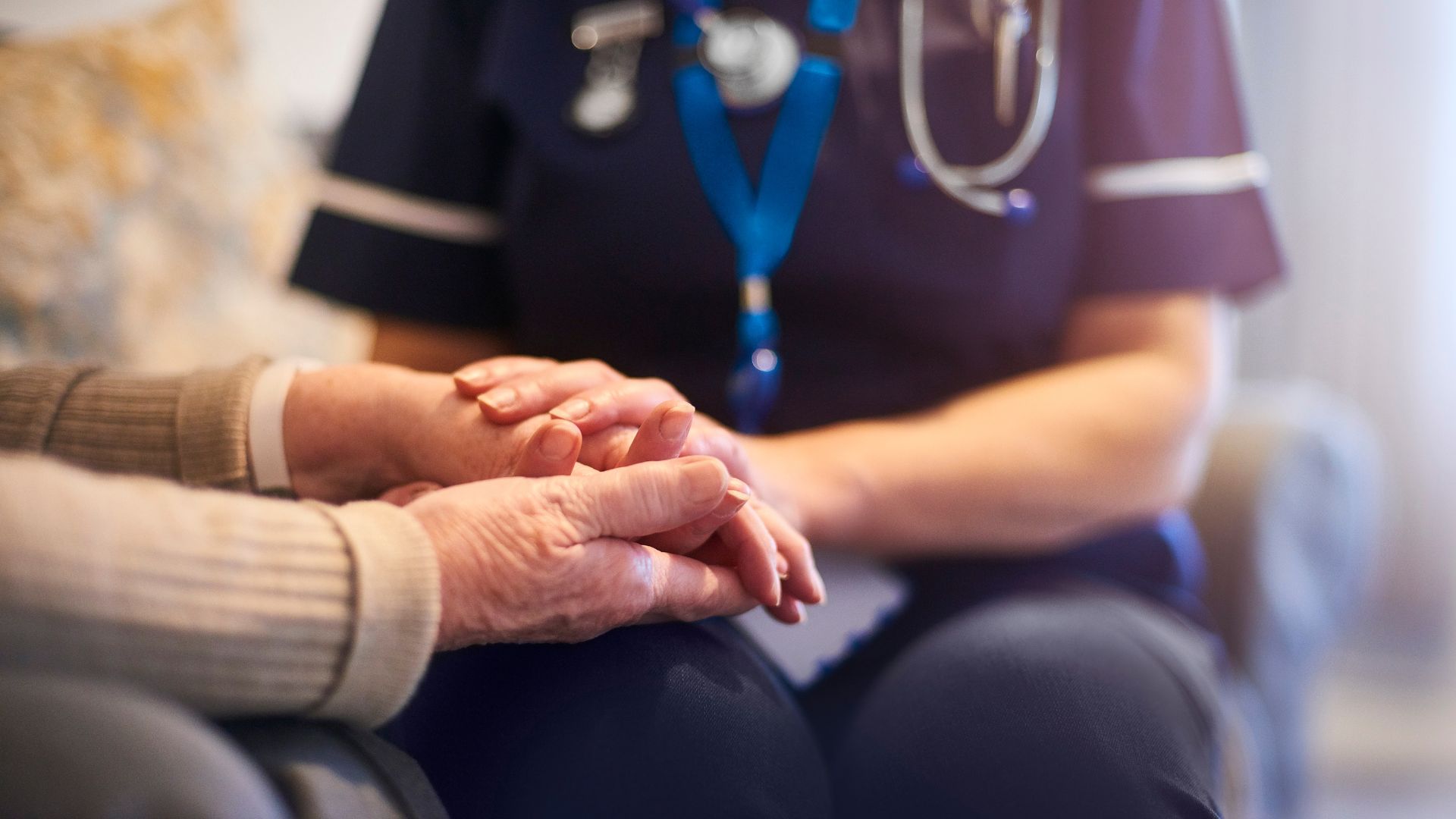 A health care worker in navy blue scrubs holds the hands of another person in a close, seated interaction..