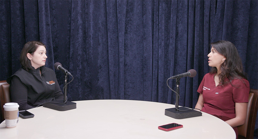 Two people sit across from each other at a round table with microphones, participating in a recorded discussion in front of a dark curtain backdrop. A coffee cup, papers, and mobile phones are visible on the table.